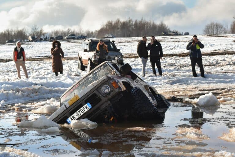 An off-road SUV is stuck in muddy snow with people standing nearby in Gjinari, Albania.