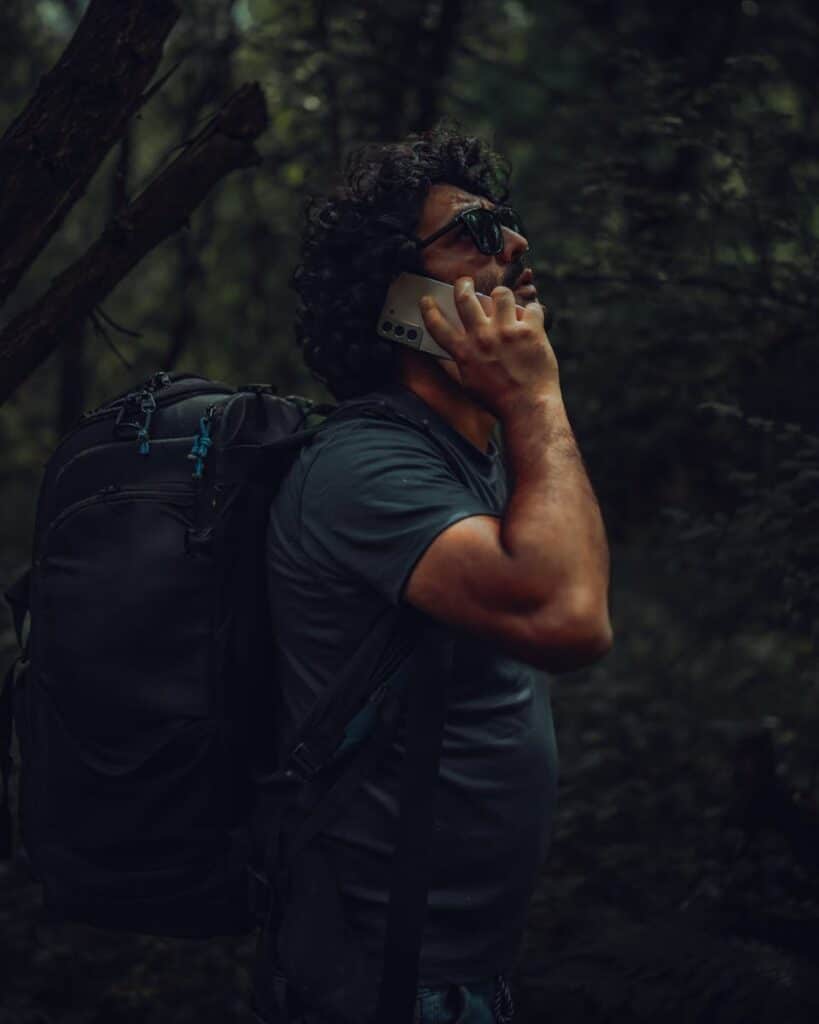 Man with backpack talking on smartphone in lush green forest setting.