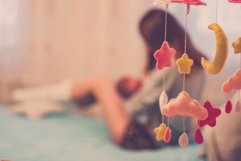 A mother cradling her baby next to a dreamy cloud and star mobile in a soft-focus bedroom setting.