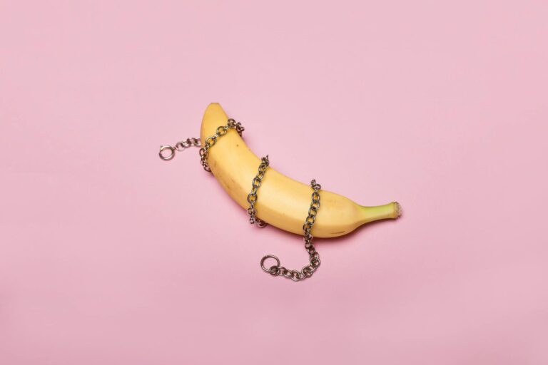 Banana wrapped in chains on pink background, symbolizing constraint and surreal art.