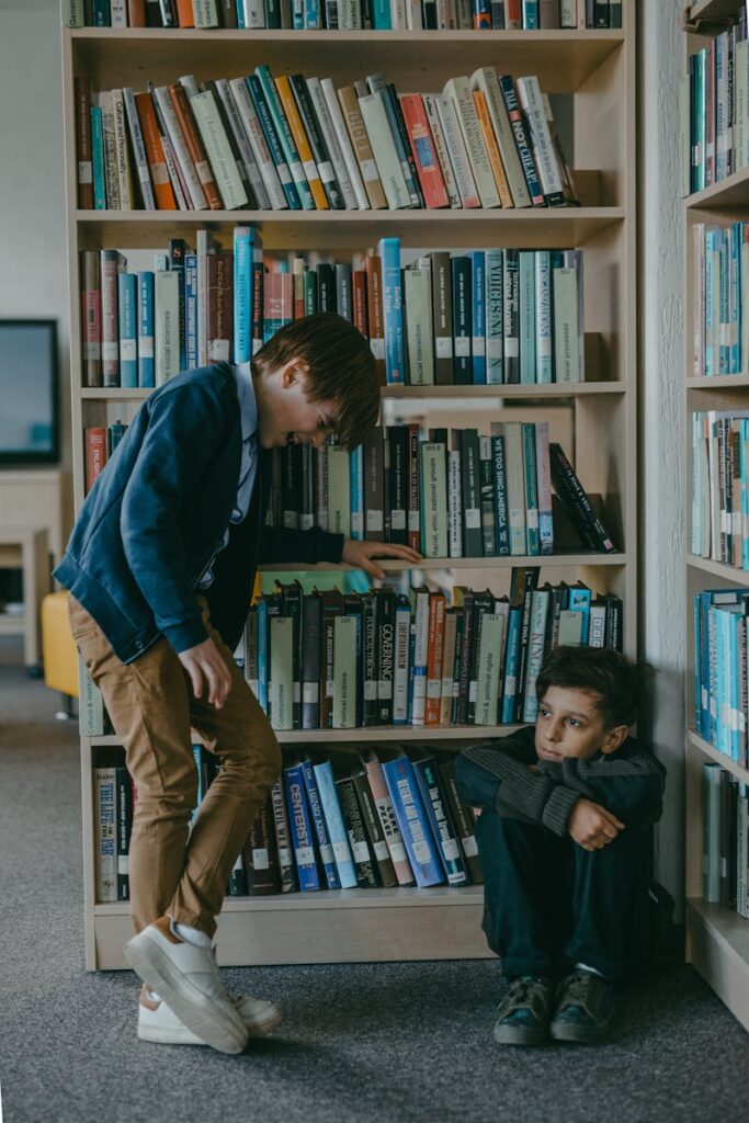 Two boys interact in a library, surrounded by bookshelves. One is sitting